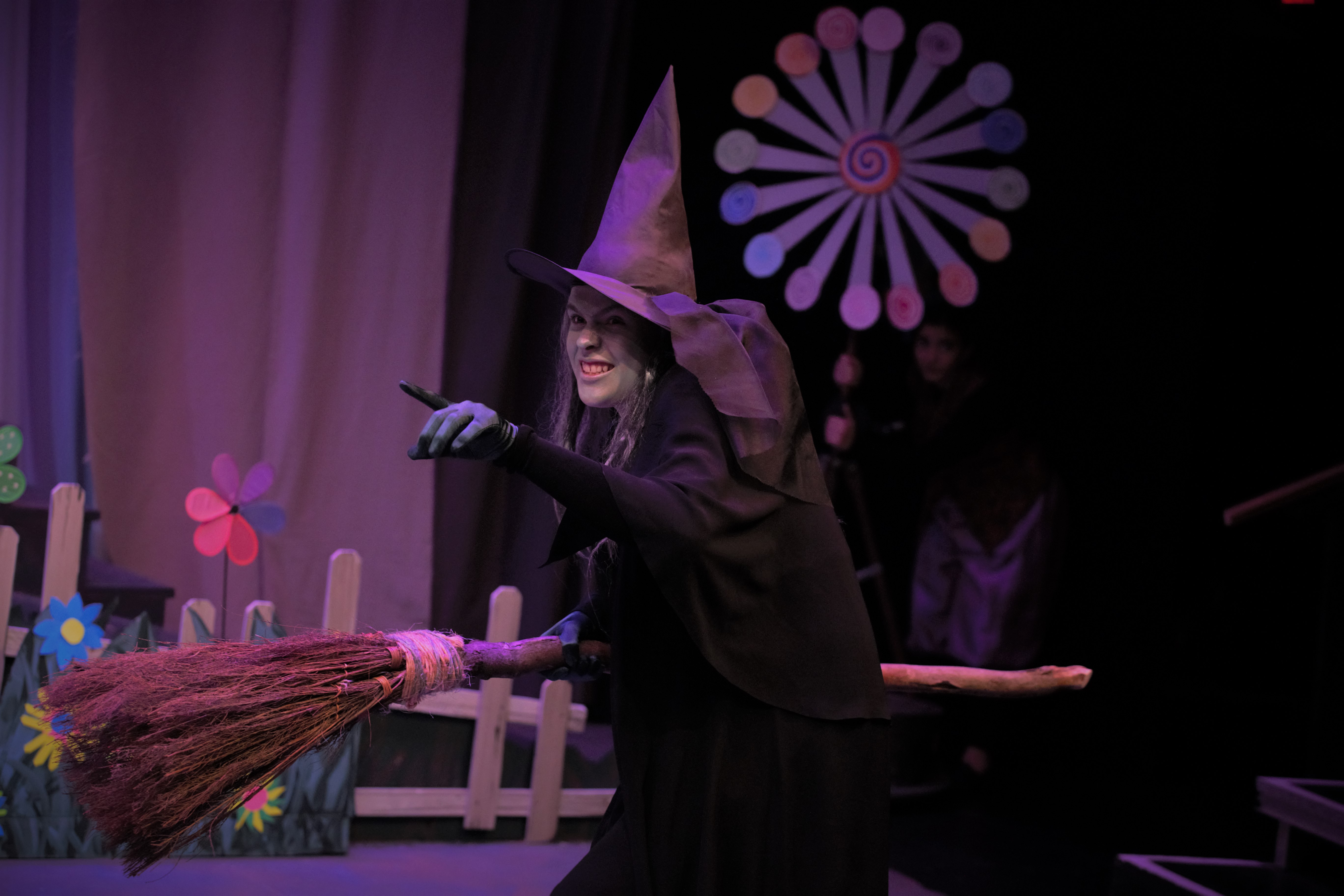 LNT student in performance of Wizard of Oz as the wicked witch of the west - green face, black dress, cape tall pointy hat, large broom in one hand - arm extended and pointing with other. 
