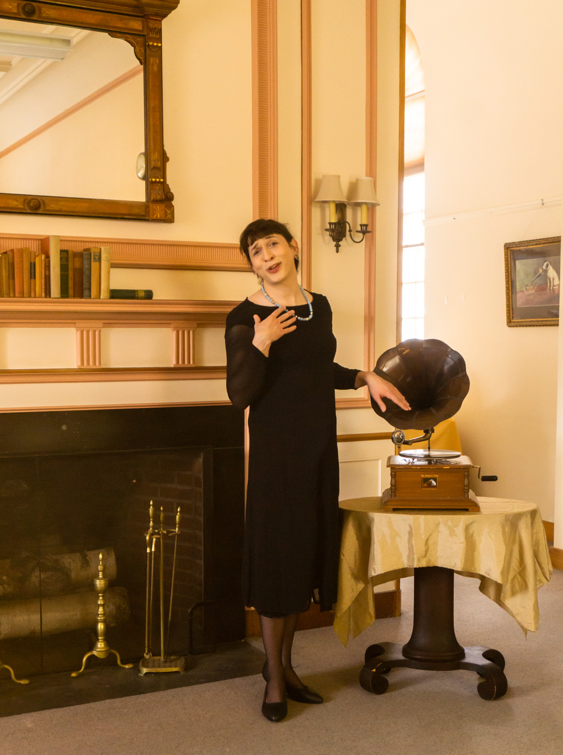 transwoman in black dress wearing blue pearl-like beads, in front of victorian fire place standing next to gramaphone